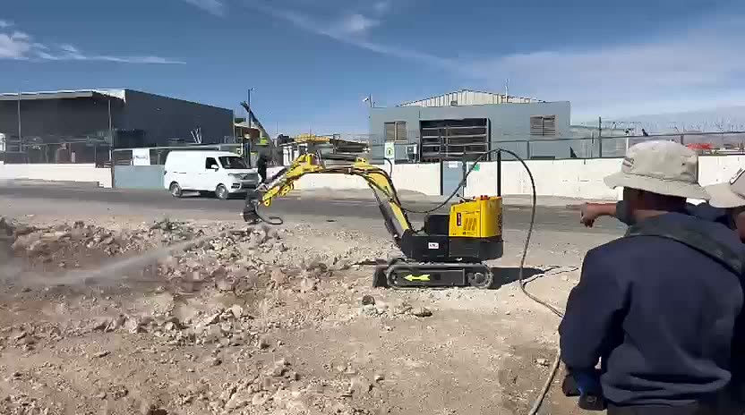 Miniexcavadora amarilla operada a distancia lanzando un chorro de agua a presión sobre material acumulado, con el equipo de trabajo observando a distancia